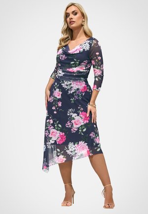 Vrouw draagt een marineblauwe midi-jurk met bloemenprint, doorschijnende driekwartmouwen en een asymmetrische zoom, poseert met handen in de zij en beige hoge hakken.