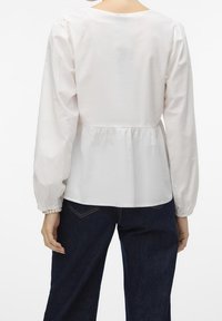 Blusa bianca a maniche lunghe con vita a peplum, scollatura rotonda e maniche arricciate. Tessuto liscio con una leggera lucentezza, abbinata a pantaloni in denim scuro.