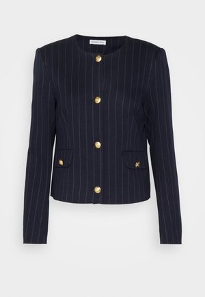 Osoba ubrana w granatową krótka marynarkę w prążki z złotymi guzikami, białą koszulę, niebieskie spodnie typu cargo i czarne, skośne buty, stojąca na tle szarej ściany.