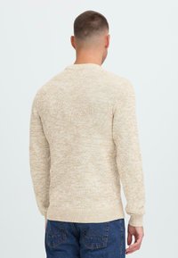 Man met kort haar in een beige gebreid sweatshirt en blauwe spijkerbroek, gezien van achteren tegen een eenvoudige witte achtergrond.