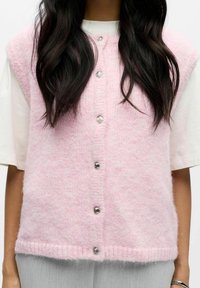 Rozā adīta veste ar mīkstu, pūkainu tekstūru, ar pogu kloķi priekšā, pieciem metāliskiem pogām un bez piedurknēm.