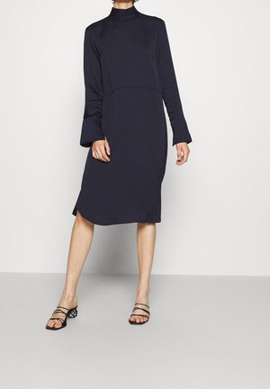 Frau trägt ein marineblaues Midi-Kleid mit langen Ärmeln und hohem Kragen sowie schwarze, offene Sandalen mit Absatz, steht vor einem schlichten Hintergrund.