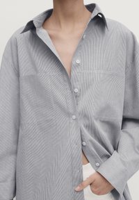 Zbliżenie na osobę ubraną w jasnoniebieską koszulę w paski z jednym ręką w kieszeni, koszula częściowo rozpięta, a kołnierz otwarty.