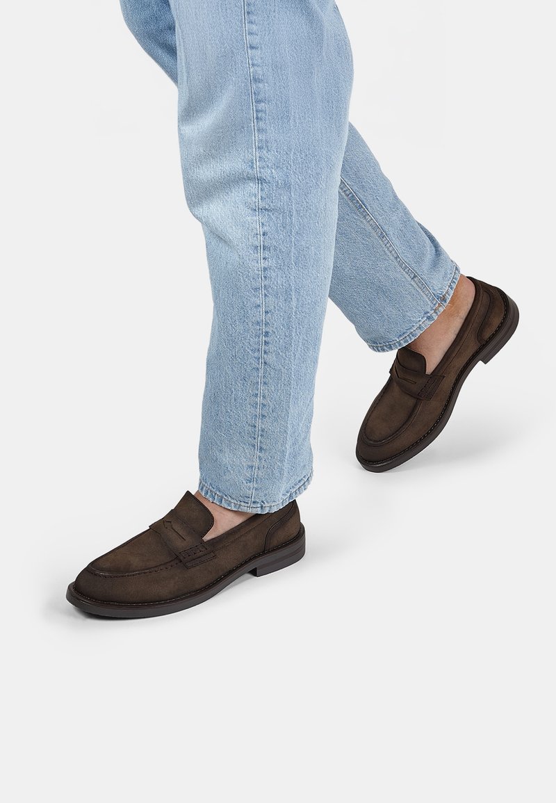 Mocasines de ante marrón con una punta redondeada, parte superior texturizada y suela de goma flexible, combinados con jeans de un azul claro en un fondo neutro.