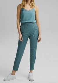 Camisole en satin teal avec dentelle, associée à un pantalon tapered teal avec une texture lisse et des plis sur le devant. Des baskets blanches complètent la tenue.