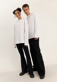 To personer iført hvide langærmede skjorter og sorte bukser står side om side mod en ensartet lys baggrund.