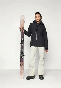 Attrezzatura da sci che include una giacca nera con zip e pantaloni grigi chiari, mentre si tiene degli sci marroni e grigi con attacchi su uno sfondo semplice.