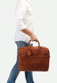 Borsa a spalla in pelle marrone con doppi manici superiori, tasca frontale e etichetta per bagaglio. Texture liscia, forma rettangolare e design minimalista.