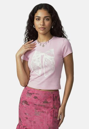 Ung kvinde med langt krøllet hår iført en lyserød crop top med hvidt blonden sløjfeprint og et lyserødt mønstret skørt med sidebind.