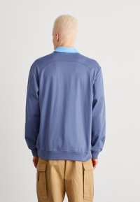 Modrá mikina se světle modrým límcem, kulatým výstřihem, dlouhými rukávy, hladkou texturou a žebrovaným lemem, viděná ze zad.