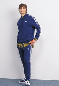 Zila Nike kapuce ar balto logo, kombinēta ar atbilstošām sporta biksēm ar sānu svītrām. Uzvilkta virs dzeltenas un melnas rakstainas krekla. Redzami sporta apavi.