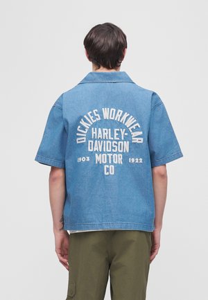Muž nosiaci modrú krátkym rukávom denimovú košeľu s textom "Dickies Workwear Harley-Davidson Motor Co 1903-1922" na chrbte.