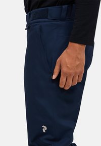 Navyblauwe broek met een gladde, rekbare textuur, voorzien van een zijrits zak en een logo nabij de zoom. Ontworpen voor comfort en mobiliteit.