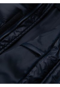 Acercamiento de una chaqueta acolchada azul marino con un bolsillo con cremallera y texturas de tela lisas y brillantes.