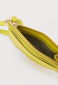 Petit portefeuille en cuir vert citron avec une fermeture éclair dorée partiellement ouverte, montrant l'intérieur vide et une sangle pour poignet sur fond blanc.