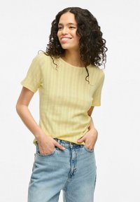 Top amarillo de manga corta con un patrón texturizado, que presenta rayas verticales y un escote redondo. Combinado con jeans de tiro alto en azul claro.