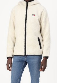 Person som bär en krämfärgad fleecejacka med dragkedja och svart trim samt "Tommy Jeans"-logotyp, ihop med blå jeans.