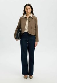 Giacca di suede marrone con colletto chiaro, camicia beige, jeans dritti blu scuro, tiene una borsa di pelle nera, indossa scarpe beige a punta.