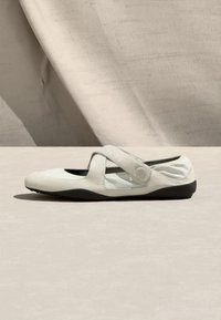 Ελαφρώς μπεζ γυναικείο παπούτσι τύπου Mary Jane με επίπεδη σόλα, ρυθμιζόμενο λουράκι, απαλό υφασμάτινο πάνω μέρος και μαύρη ευέλικτη σόλα σε ουδέτερο φόντο.