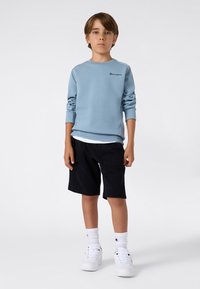 Un jeune garçon se tient debout, portant un sweat-shirt bleu clair, un short noir, des chaussettes blanches et des sneakers blancs, contre un fond uni clair.