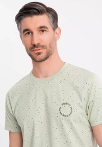 Lichtgroene t-shirt met korte mouwen, voorzien van een gespikkeld patroon en een ronde afbeelding met de tekst: "jouw enige beperking is je geest."
