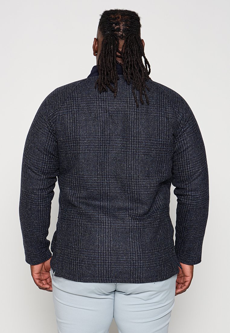 Mann mit langen Dreadlocks, der eine dunkelblaue karierte Jacke und hellgraue Hosen trägt, steht mit dem Rücken zu einer weißen Hintergrund.