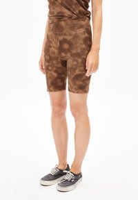 Shorts de vélo marron tie-dye avec une texture de tissu lisse et une taille haute, associés à des baskets noires. Les détails incluent un petit logo sur la ceinture.