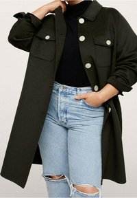 Chaqueta de gran tamaño en color verde olivo con botones grandes y dos bolsillos en el pecho, combinada con jeans rotos de color azul claro. El tejido parece texturizado.