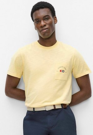 Homme à la peau foncée portant un t-shirt jaune pâle avec un petit logo sur la poche, un pantalon bleu marine et une ceinture tissée beige, debout contre un mur blanc.