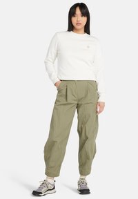 Krämfärgad tröja med rund halsringning och liten logotyp; ljus olivgrön, veckad, tapered byxor; grå och lila vandringsskor.