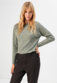 Blonde woman with long hair wearing a light gray crewneck sweater and dark brown trousers, posing with one hand on hip.