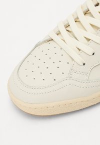 Beige lærresneaker med rund tå, perforerte detaljer, flate kremfargede snørebånd og teksturert gummisåle. Sidesømmer gir dimensjon.