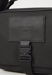 Sac noir texturé avec un patch en cuir étiqueté "Pier One Berlin" et une boucle métallique noire attachée à une sangle.