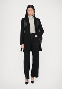 Fekete hosszú kabát bőrt részletekkel és rojtos díszítéssel, csíkos garbó felett viselve. Széles szárú fekete nadrággal és magassarkú cipővel párosítva.