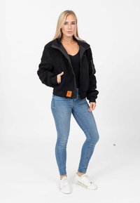 Giacca bomber in pile nera con zip, polsini elasticizzati, tasche laterali e un'etichetta arancione. Indossata con jeans blu e scarpe da ginnastica bianche.