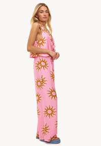 Combinaison rose avec des motifs de soleil en orange. Design sans manches, coupe décontractée, taille élastique, pantalons longs et fentes latérales ; bordures unies aux bords.