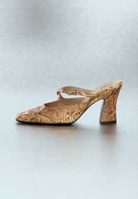 Καφέ mule με σχέδιο δέρματος φίδι, τετράγωνη μύτη, σταυρωτό λουρί και χοντρό τακούνι σε ανοιχτό γκρι φόντο.