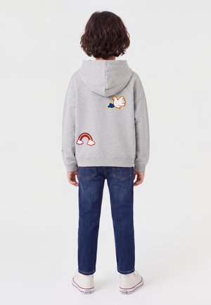 Enfant aux cheveux bouclés vu de dos portant un sweat à capuche gris avec des patches colombe et arc-en-ciel, un jean bleu foncé et des baskets blanches.