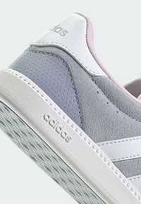 Šedá semišová a kožená teniska s bielymi akcentmi, s textúrovaným povrchom, zaokrúhlenou špičkou a vzorovanou gumovou podrážkou. Viditeľné označenie značky „Adidas“.