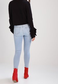 Svart kort tröja med volanger på ärmarna, kombinerad med ljusblå skinny jeans och röda ankelstövlar. Outfiten visas från en bakvy.