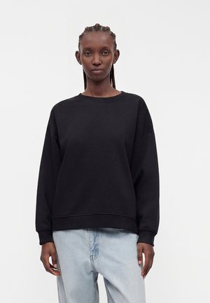 Zwarte crewneck sweatshirt gedragen door een staand persoon met gevlochten haar en lichtblauwe spijkerbroek tegen een effen witte achtergrond.