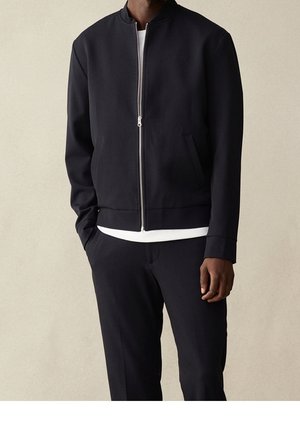 Mann trägt eine schwarze Reißverschlussjacke über einem weißen Hemd mit schwarzer Hose, steht vor einem einfarbigen beigen Hintergrund.