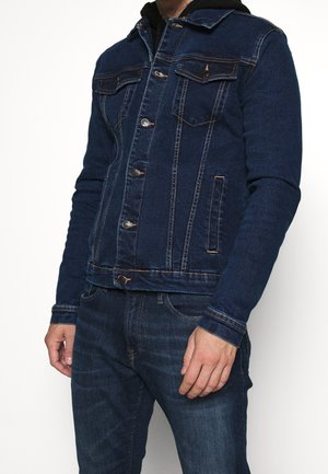 Mężczyzna ubrany w ciemnoniebieską kurtkę dżinsową i dżinsy z zapięciem na guziki z przodu, stojący na jednolitym białym tle.