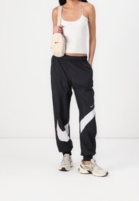 Czarne sportowe spodnie dresowe z białym logo Nike i paskiem, zestawione z jasnoszarym tank topem i beżową torebką na ramię. Buty sportowe w neutralnym kolorze.