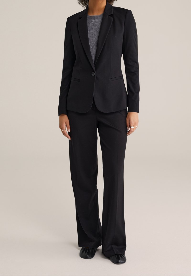 Femme portant un blazer noir ajusté, un pantalon large assorti, un haut en maille gris, des chaussures noires et des bagues argentées sur fond beige.