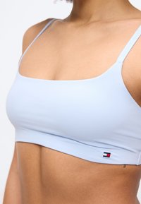 Γυναίκα που φοράει αθλητικό σουτιέν ανοιχτού γαλάζιου χρώματος με λεπτές τιράντες και μικρό λογότυπο Tommy Hilfiger στην κάτω πλευρά.