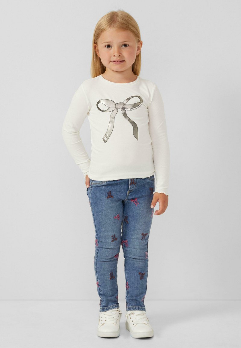 Witte langslavende shirt met een zilveren strik graphic, gecombineerd met blauwe jeans met roze strik borduursel en witte sneakers.