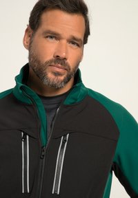 Svart och grön zip-up jacka med hög krage, som har två bröstfickor med kontrasterande dragkedjor och en texturerad tyg.