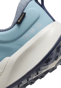 Chaussure bleu clair avec un dessus en tissu texturé et une semelle intermédiaire blanche. Présente un logo et trois symboles sur le côté, avec une semelle extérieure en caoutchouc foncé.