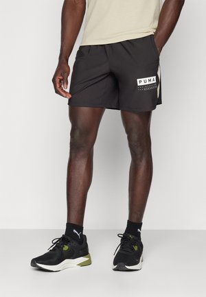 Mann trägt schwarze Puma-Sportshorts, schwarze Puma-Socken und schwarz-weiße Laufschuhe vor einfachem Hintergrund.